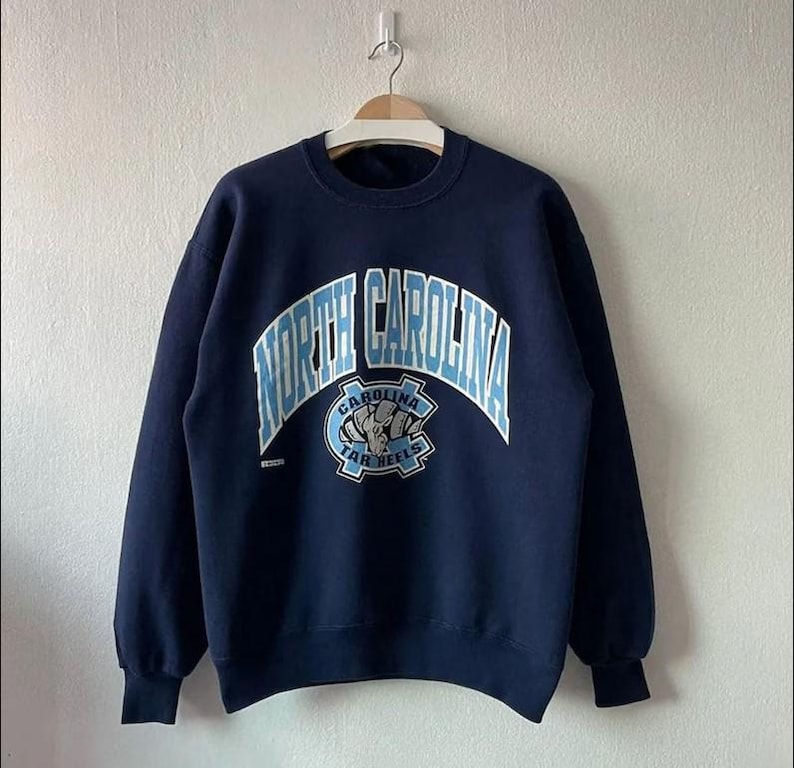Puede incluir: Sudadera azul marino con la inscripci&oacute;n "NORTH CAROLINA" en azul claro arqueada sobre el pecho. Debajo, un logotipo azul claro con "CAROLINA TAR HEELS". La sudadera est&aacute; colgada de una percha de madera.