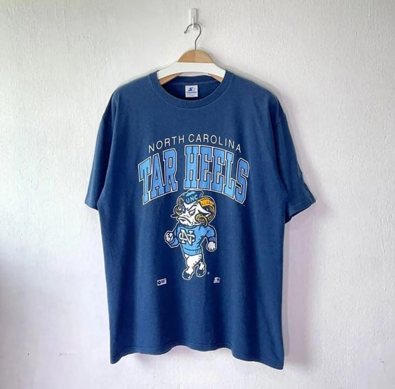 May include: A navy blue t-shirt featuring the words "North Carolina Tar Heels" in light blue arched lettering. Below the text is a cartoon ram mascot in a light blue uniform. The shirt is hanging on a white wall.