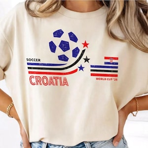 May include: A cream-colored t-shirt featuring a distressed graphic of a blue soccer ball, three stars, and horizontal stripes in blue, black, and red. The text reads SOCCER CROATIA and WORLD CUP '26, with a small Croatian flag emblem included.