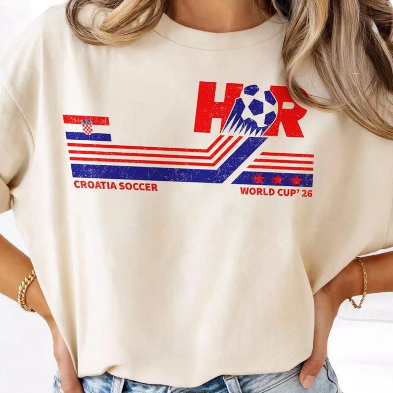 May include: Cream-colored t-shirt featuring a graphic design with the text "HOR" and a soccer ball. The design incorporates the Croatian flag colors: red, white, and blue. The text "CROATIA SOCCER" and "WORLD CUP '26" are also included.