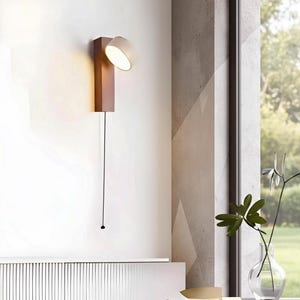 May include: A modern wall-mounted lamp with a rectangular brown base and a circular, adjustable light head. The lamp is attached to the wall with a long black cord and a small pull switch. The light is on, illuminating the surrounding area.