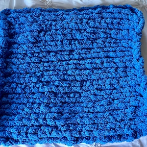 May include: A square, hand-knitted blanket in a vibrant shade of blue. The blanket features a chunky knit texture, with visible loops and a soft, plush appearance. The edges are neatly finished, creating a cozy and inviting aesthetic.