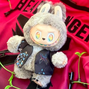 May include: A plush toy with gray fur, rabbit ears, and rainbow-colored eyes wearing a black vest with colorful floral and geometric patterns. The toy is positioned on a bright red fabric background featuring black text and green sprout clips.