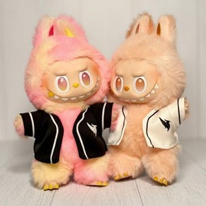 May include: Two plush bunny figures with expressive faces and furry bodies. One is pink and yellow, the other is peach. Both wear jackets with a white bird logo. The figures have large eyes and small, sharp teeth.