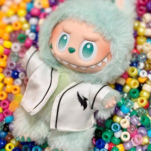 May include: A plush toy with a light green furry hood and matching body. The toy has a white jacket with black trim and a black bird logo. The background is a colorful assortment of beads.