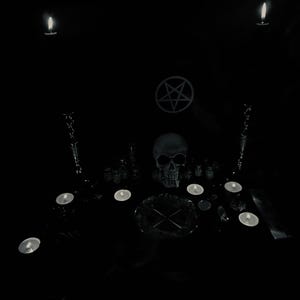 May include: A dark, moody scene featuring a human skull, lit candles, and a pentagram symbol. The arrangement includes various ritualistic objects, creating a gothic or occult aesthetic. The image is predominantly black and white.