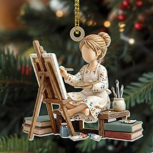 May include: A Christmas ornament depicting an artist painting on a canvas. The artist is seated on a small stool, wearing a floral-patterned apron and holding a palette. Paint tubes, brushes, and books are also present. The ornament is suspended by a gold string.