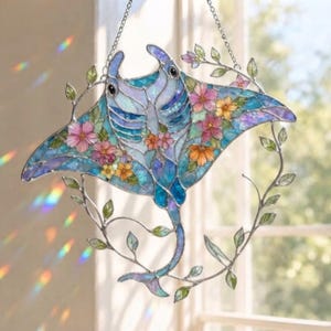May include: A stained glass suncatcher shaped like a manta ray, featuring iridescent blue and purple glass panels. The manta ray is decorated with pink and yellow floral accents and surrounded by a delicate metal vine frame with green leaves.