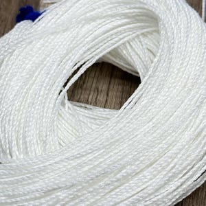 May include: A tightly wound skein of white cord, likely yarn or string, is the central focus. The cord appears to be made of a smooth, possibly synthetic material. The background is a wooden surface. A small amount of blue material is visible at the top.