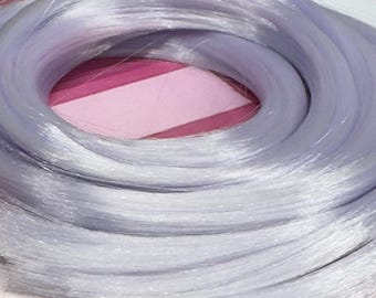 May include: A coiled bundle of light purple synthetic hair extensions. The hair is a pale lavender color and appears shiny. The bundle is tied with a small piece of green material. The background is pink and white.