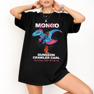 Op de afbeelding: Zwart t-shirt met het woord "MONGO" boven een blauwe en roze dinosaurusillustratie. Onder de illustratie staat de tekst "DUNGEON CRAWLER CARL" en Japanse karakters. Een casual kledingstuk.