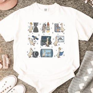 Op de afbeelding: Wit T-shirt met een grillig ontwerp van cartoon ganzen die zich bezighouden met ruimtevaartactiviteiten. De illustraties omvatten een raket, een astronaut, een satelliet en bedieningspanelen. De ganzen dragen helmen en andere ruimtevaartuitrusting.