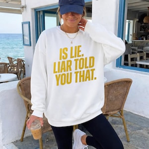 Può includere: Felpa bianca con la scritta gialla "IS LIE. LIAR TOLD YOU THAT.". La felpa è a maniche lunghe e ha il girocollo. La persona nell'immagine indossa un berretto da baseball blu navy e leggings neri.