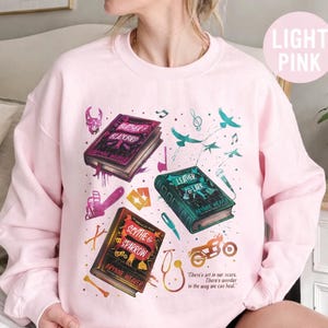 May include: Light pink sweatshirt featuring book illustrations with the titles "Butcher & Blackbird", "Leather & Lark", and "Scythe & Sparrow". The design includes musical notes, birds, and the quote "There's art in our scars. There's wonder in the way we can heal."