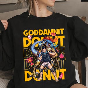 May include: Black sweatshirt with a graphic design featuring the text "GODDAMNIT DONUT" in yellow. The design includes a cartoon dinosaur, a man, and various donut-themed illustrations. The sweatshirt has long sleeves.