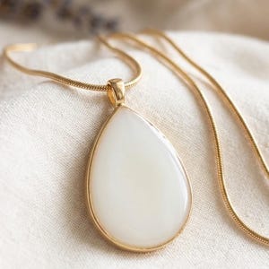 Può includere: Una collana con pendente a goccia caratterizzata da una pietra bianca opaca e liscia incastonata in una cornice di metallo dorato lucido. Il pendente è appeso a una catena a serpente dorata abbinata, creando un gioiello pulito ed essenziale.