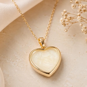 Puede incluir: Un collar dorado con un colgante en forma de corazón. El colgante tiene un centro blanco cremoso y un borde dorado. El collar está sobre una superficie de color claro.