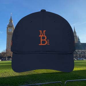 May include: Navy blue baseball cap with an embroidered orange design. The design features the letters "MB" with "ond" below. The cap has a curved brim and a structured crown. The background shows a famous clock tower and a green lawn.