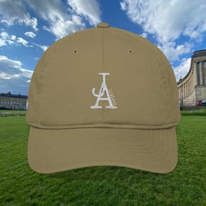 May include: A khaki baseball cap with a curved brim. The cap features a white embroidered design of the letter "A" with the word "listen" vertically along the side. The cap is set against a backdrop of a green field and a cloudy sky.