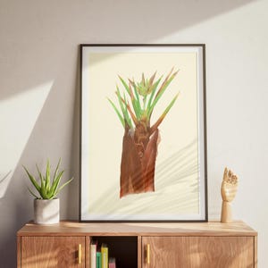 Puede incluir: Una impresión botánica enmarcada con una ilustración en acuarela de una planta con tonos marrones, verdes y naranjas. La obra de arte se exhibe sobre un fondo beige claro, enmarcada en negro, sobre un mueble de madera.