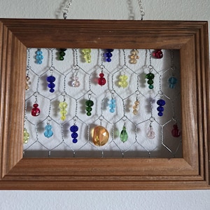 May include: A rectangular wooden frame containing a silver wire mesh grid. Hanging from the wire mesh are numerous small, multi-colored glass beads in shades of red, blue, green, yellow, and pink. A silver chain is attached to the top for hanging.