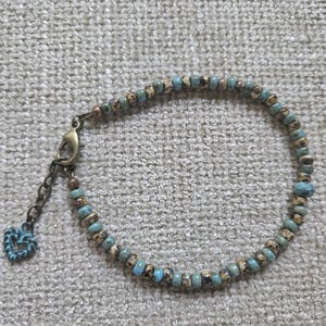 May include: A beaded bracelet featuring small, round, turquoise-blue and speckled tan beads arranged in a repeating pattern. The bracelet includes a bronze-toned metal lobster clasp, an adjustable chain link, and a small heart-shaped charm.