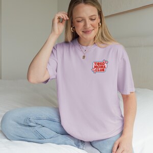 May include: A light purple t-shirt with the text "TIRED MOMS CLUB" in a red and blue design. The shirt is worn with light blue jeans. The image shows a person wearing the shirt.