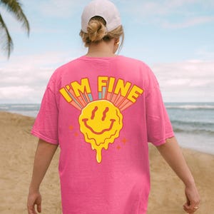 May include: A bright pink t-shirt with the words "I'M FINE" above a yellow melting smiley face. The design has retro-style colors and is printed on the back of the shirt. The person is wearing a white cap.