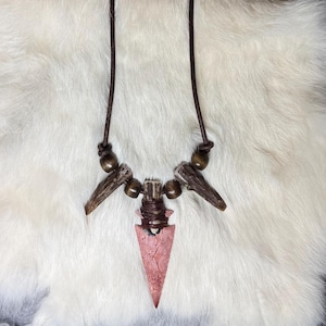 May include: A handcrafted necklace featuring a pink arrowhead pendant, antler accents, and wooden beads. The necklace has a brown leather cord with adjustable knots. The background is a white, fluffy surface.