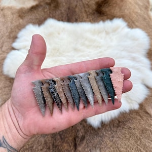 May include: A hand holds fifteen stone arrowheads in a variety of colours, including grey, brown, and green. The arrowheads have serrated edges. They are displayed against a backdrop of a fur rug.