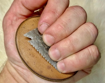 May include: A round, brown leather object with a serrated, gray stone blade. The leather has a stitched border. The stone blade is triangular and has a rough, textured surface. The object is held in a hand.