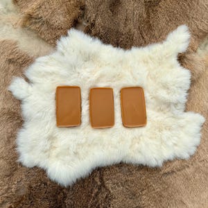 May include: Three caramel-coloured rectangular objects are displayed on a white, fluffy surface, with a brown fur background. The objects have rounded corners and a glossy finish, possibly made of resin or a similar material.