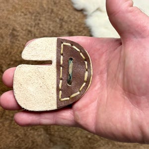 May include: A leather archery finger tab, designed to protect the fingers. The tab is a combination of light tan and brown leather, stitched together with cream-colored thread. A green and brown woven material is visible in the center.