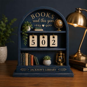 May include: A dark blue arched wooden bookshelf display with three shelves. The top shelf features a globe, a small potted plant, and the text BOOKS read this year. The middle shelf holds three numbered tags reading 502. The bottom shelf contains miniature books, a succulent, and a lantern, with the text JACKSON'S LIBRARY printed on the base.