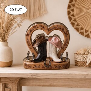 May include: A wooden heart-shaped display featuring a brown cowboy hat on a black boot and a pink cowboy hat on a white boot. The base is engraved with the names Jack and Emily, and the heart frame reads Together We Ride, Forever Side By Side.