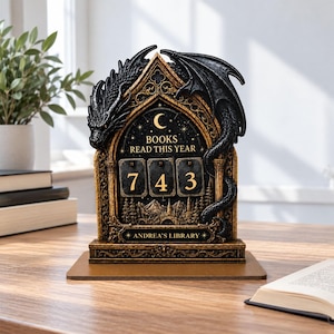 May include: A black and gold book tracker featuring a sculpted dragon perched atop a gothic archway. The center displays the text BOOKS READ THIS YEAR above three rotating number tiles showing 743 and the name ANDREA'S LIBRARY below.