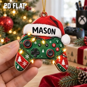 May include: A flat, two-dimensional Christmas ornament shaped like a video game controller. The controller is green with red accents, decorated with white snowflakes and trees. It features a Santa hat with the name MASON and string lights.