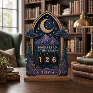 May include: A wooden arched desk display featuring a night sky with a crescent moon and stars above purple mountains and evergreen trees. It includes the text "BOOKS READ THIS YEAR" above three black flip-style number tiles showing 126 and a nameplate reading "OLIVIA".