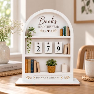 May include: A white arched wooden bookshelf display with three tiers. The top shelf features the text Books READ THIS YEAR with a heart icon, miniature books, and a hanging plant. The middle shelf displays flip cards showing 279. The bottom shelf holds miniature books, a small potted plant, and a candle. The base is labeled DIANNA'S LIBRARY.