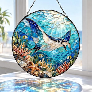 May include: A circular stained glass suncatcher featuring a manta ray swimming through blue ocean water. The scene includes coral reefs, a starfish, and small yellow and black fish. The piece is framed in black metal and hangs from a chain.