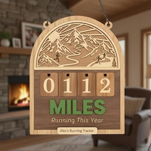 May include: Wooden running tracker with a mountain scene and the text "0112 MILES Running This Year." The tracker has a chain for hanging and a space for a name. The design features a sun, mountains, and runners.
