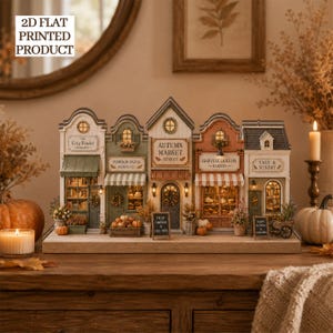 May include: A 2D flat printed product featuring a row of five miniature autumn-themed storefronts including The Cozy Reader Bookshop, Pumpkin Patch Floral Co., Autumn Market Street, Harvest Hollow Bakery, and Sage & Sundry on a wooden base.
