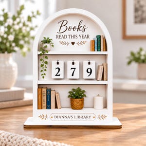 May include: A white arched wooden bookshelf display with three tiers. The top shelf features the text "Books READ THIS YEAR" with a heart icon, miniature books, and a hanging plant. The middle shelf has flip-style number cards showing 279. The bottom shelf displays miniature books, a small potted plant, and a white candle, with the text "DIANNA'S LIBRARY" printed at the base.