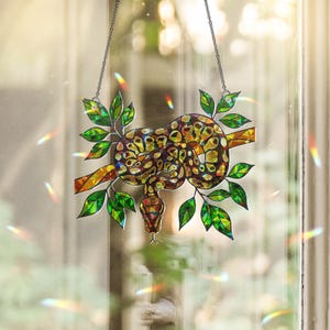 Op de afbeelding: Gebrandschilderd glazen raamhanger met een gedetailleerde slang op een tak met groene bladeren. De slang heeft een bruin en geel patroon. Het kunstwerk wordt opgehangen met een ketting, waardoor een decoratieve zonnevanger ontstaat.