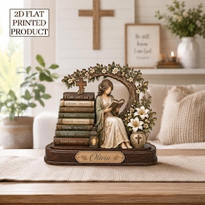 May include: A 2D flat printed decorative figurine featuring a person reading a book beside a stack of seven books labeled with religious phrases. The scene includes a tree arch, white lilies, a small lantern, and a nameplate reading Olivia.