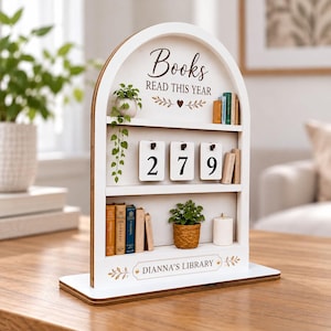 May include: A white arched wooden bookshelf display with three tiers. The top features the text "Books READ THIS YEAR" with a heart icon. The middle tier has three flip cards showing the number 279. The bottom tier displays "DIANNA'S LIBRARY" with small potted plants, miniature books, and a candle.