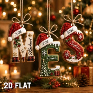 May include: Three hanging Christmas ornaments shaped like the letters M, E, and S. Each letter features a red Santa hat, the year 2026, and a personalized name: Michael, Emma, and Sophia. The ornaments are decorated with snowflakes, pine, and gifts.