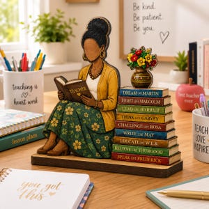 Puede incluir: Un adorno de escritorio de madera con una figura de peinado alto, vistiendo un c&aacute;rdigan amarillo y una falda verde con flores, leyendo un libro que dice TEACHER. Junto a la figura hay una pila de libros con nombres como MARTIN y MALCOLM.