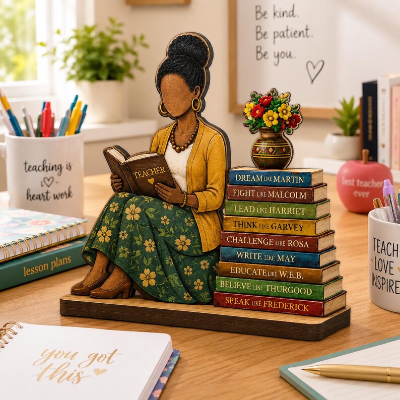 Puede incluir: Un adorno de escritorio de madera con una figura de peinado alto, vistiendo un c&aacute;rdigan amarillo y una falda verde con flores, leyendo un libro que dice TEACHER. Junto a la figura hay una pila de libros con nombres como MARTIN y MALCOLM.