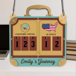 May include: A suitcase-shaped travel sign with a gold handle and teal border. The sign has a tan center with the text "Countries Visited" and "US States", along with number counters. The sign also displays the text "Emily's Journey". The sign is designed to be hung.